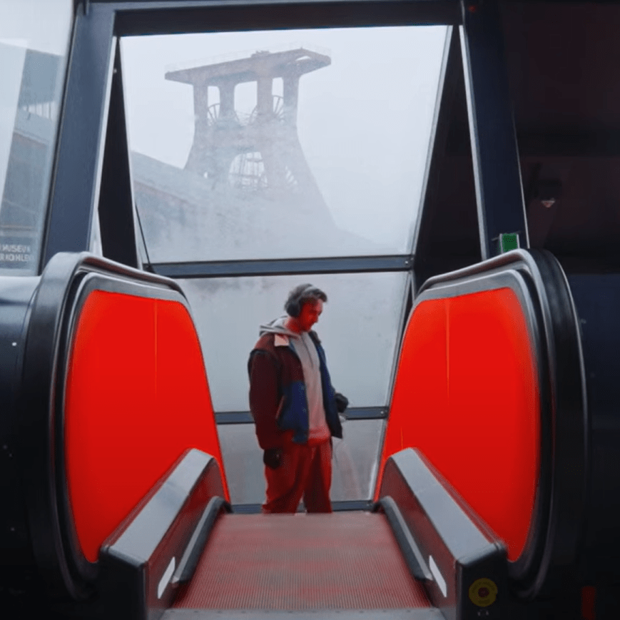 Nils Mosh on an escalator , recording with a contact mic and the mining tower of Zeche Zollverein in the background